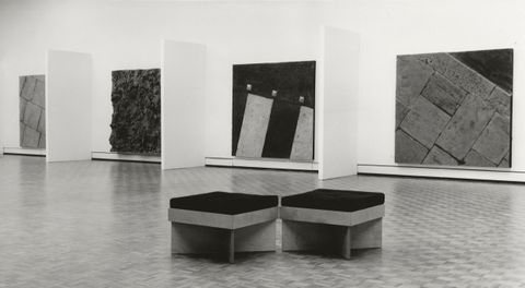 Installation photograph. Black and white photograph of four panels hung on a gallery wall. Each is square and is a cast of different kinds of ground, like pavers or a patch of dirt. Short projecting walls create barriers between them.