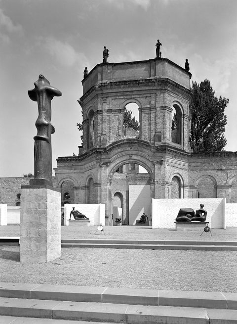 Installation photograph. Black and white photograph shows sculptures displayed against white walls set up in front of the remains of a stone building. Most of the sculptures are on plinths in this view, and the few near the central, arched doorway of the building are of stylized human bodies.