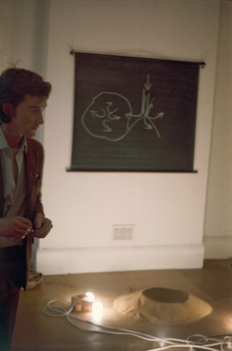 Colour photograph. A pale-skinned man with dark hair stands to our left, almost out of frame. A black picture with a drawing or diagram in white hangs on the wall behind him. Two lights lie on the floor next to a tan-coloured mound.