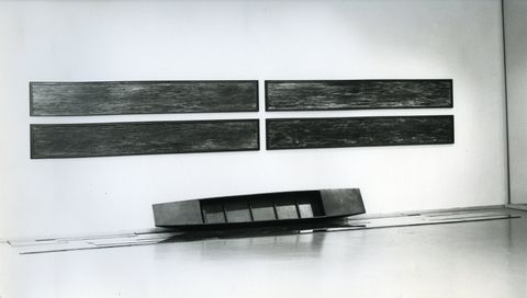 Installation photograph. Black and white photograph of a long, narrow boat propped longways against a gallery wall. Four equally long, textured panels hang above, two by two.