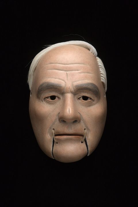 Sculpture. Wooden mask of a man’s face has pale skin, white hair, and wrinkles across the forehead. Hooded eyes look out under heavy black brows. The moveable chin is tied onto the mouth with twine.