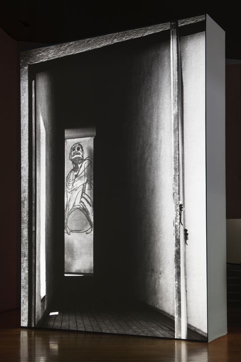 Installation photograph. A black and white film is projected onto a floating wall. This still shows a tall, narrow drawing seen at the end of a hall. The drawing is of a stylized person with a skull-like face.
